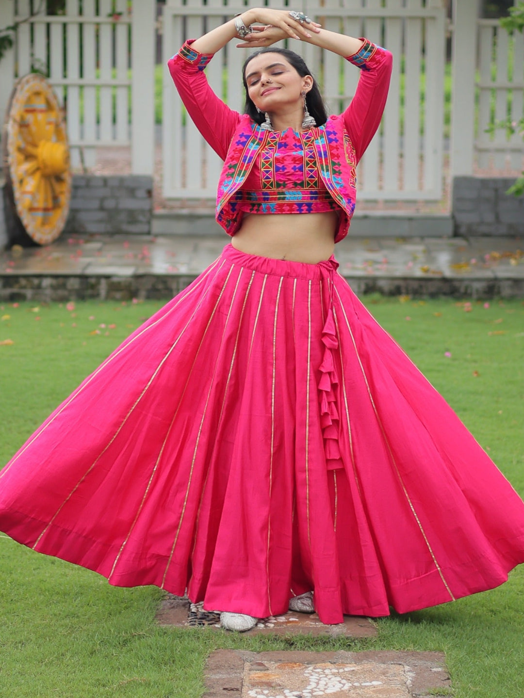 Woman in a bright pink traditional outfit with a colorful top, standing on grass with a decorative archway in the background.
