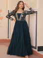 Woman in a dark blue dress with floral embroidery standing in an ornate room.