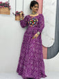 Woman wearing a purple dress with a colorful pattern, standing against a white wall.