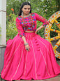 Woman in a bright pink traditional outfit with colorful blouse, sitting next to a large wheel with decorative patterns.