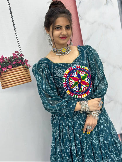 Woman wearing a green traditional outfit with a colorful embroidered design, standing against a white wall with a hanging plant.
